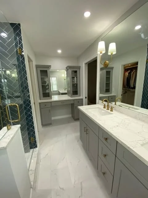 Modern bathroom with gray cabinetry, marble countertops, and a large mirror. Features a walk-in shower with dark blue tile and gold fixtures, and a walk-in closet with clothes visible.
