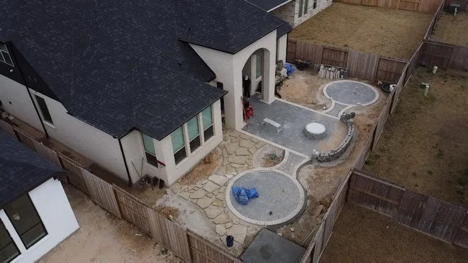 A backyard with two circular paved areas, a small round hot tub, and a stone pathway leading to the house entrance, all enclosed by a wooden fence.