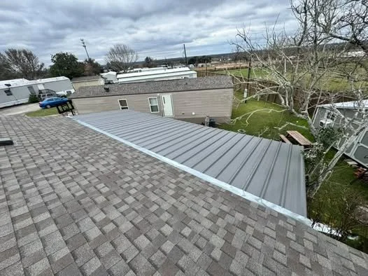 View of rooftops with shingle and metal roofing, a tree, and mobile homes or trailers in a park under a cloudy sky.