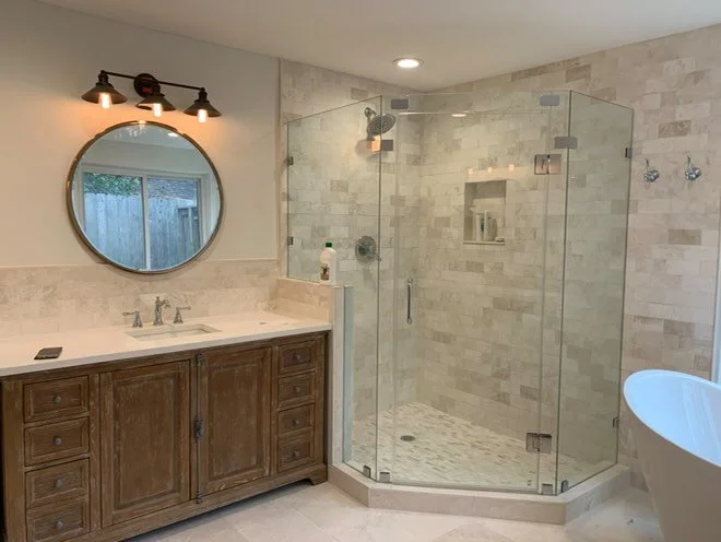 Bathroom with wooden vanity, round mirror with black light fixtures, glass-enclosed shower with beige tiled walls, small built-in shower shelf, and a free-standing bathtub.