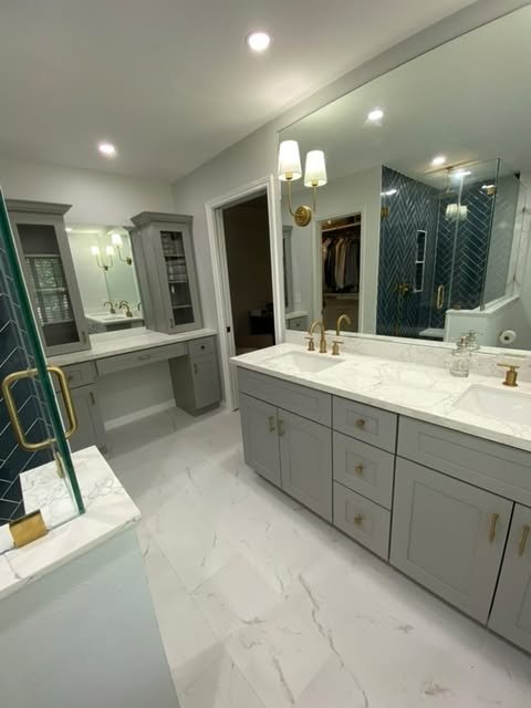 Modern bathroom with white marble countertops, gray cabinetry, and a large mirror. There is a double sink with gold fixtures, wall-mounted lights, and a glass-enclosed shower with dark blue tiles.