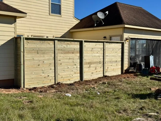 Newly built wooden privacy fence in a backyard, with a neighboring house, satellite dish, and garden tools nearby.