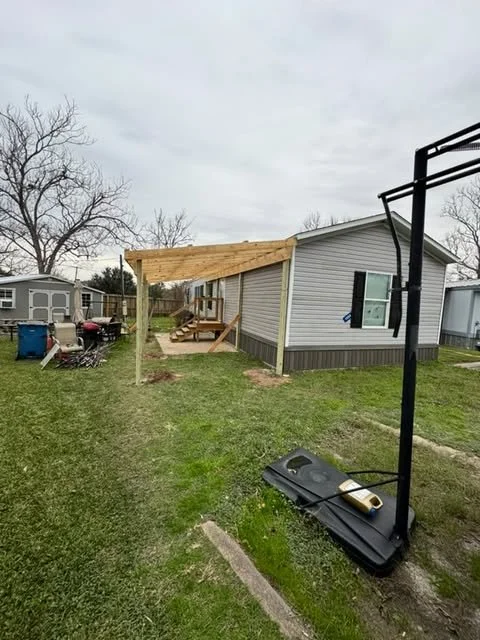 Backyard with newly constructed wooden porch extension on house, basketball hoop with broken backboard lying on the ground, and other mobile homes in background.