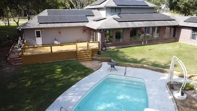 Residential backyard with a swimming pool, a hot tub, a grassy lawn, a wooden deck, and a house with solar panels on the roof.
