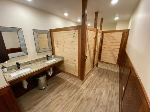 Public bathroom with two sinks, mirrors, privacy stalls, wooden partitions, and wooden paneling on the wall.