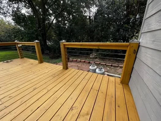 Wooden deck with cable railing and two propane tanks in the background, outdoor setting with trees and a house wall on the right.
