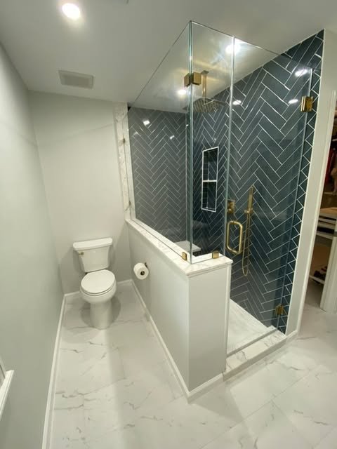 A small modern bathroom with a toilet and a glass-enclosed shower featuring dark blue herringbone tiles and gold fixtures.