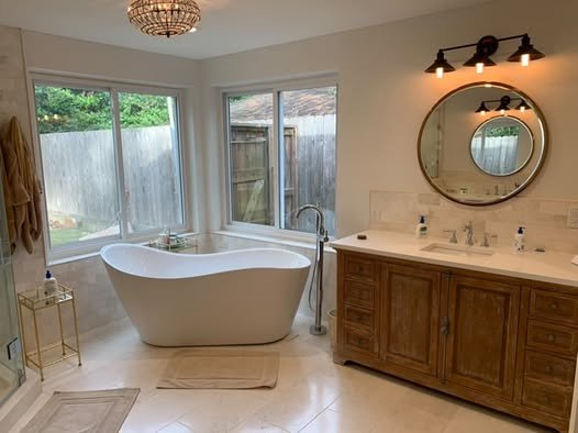 Bathroom with a white bathtub near large windows, wooden vanity with a round mirror, and a light fixture above the mirror.