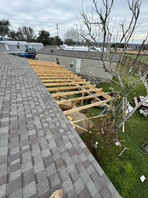 View of a house roof with gray shingles, adjacent to a construction area with wooden beams forming a new structure, a tree with no leaves, and neighboring homes in the background under a cloudy sky.
