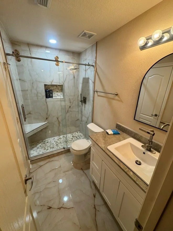 Bathroom with a glass-enclosed shower, white toilet, a white vanity with a granite countertop, a mirror, and wall-mounted lights.