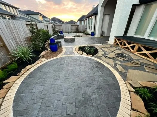 Backyard patio with pavers, plants, and blue decorative planters. Fencing and neighboring houses visible in the background at sunset.