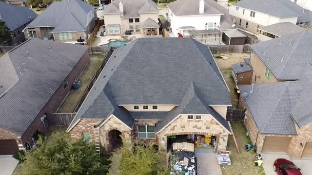 Aerial view of a large house with a dark gray roof, surrounded by neighboring houses with similar styles, and a backyard with trees and a fenced yard.