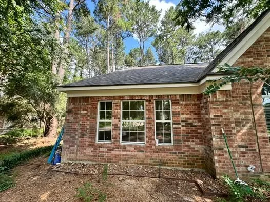 Back of a brick house surrounded by trees, with three vertical windows and gardening tools leaning against the wall.