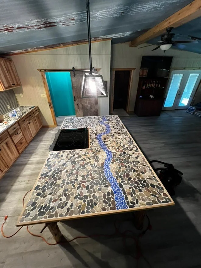 A kitchen island with a mosaic design of a blue stream running through pebbles, surrounded by wooden cabinetry and a dark ceiling with exposed beams.