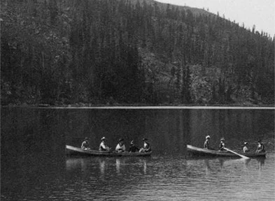 Two canoes with six people paddling on a calm lake with a forested mountain in the background.