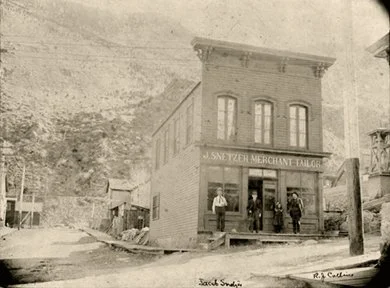 An old black and white historic photograph of a two-story building labeled 'J. Snedzer Mercantile Tailor' with three men standing in front. The building has large windows and a prominent sign. Surrounding structures are visible nearby.