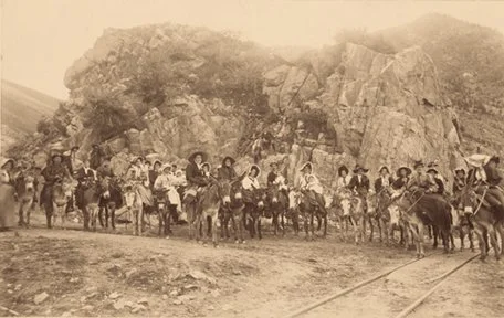 A sepia-toned historical photograph of a group of people and animals walking along a rural road with rocky hills in the background.