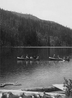 People in canoes on a calm lake with forested hills in the background.
