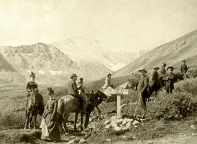 Group of people, some on horses, gathered near a sign in a mountainous outdoor area.