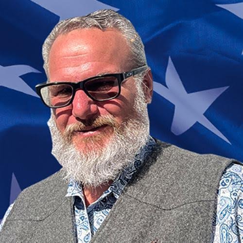 Man with gray hair and beard wearing glasses, a gray vest over a purple checkered shirt, standing in front of multiple flags including California state flags and American flags.