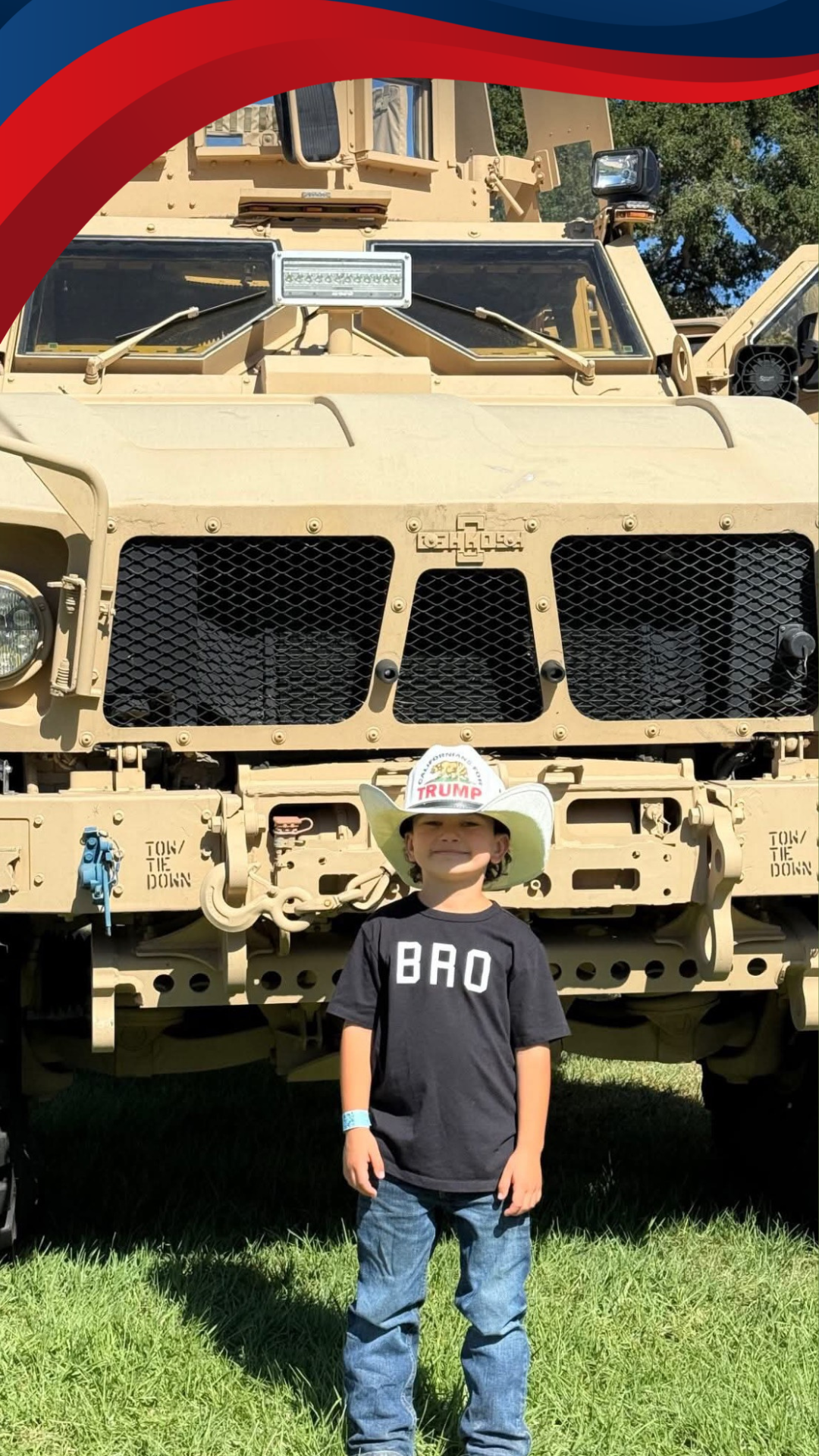 A young boy stands in front of a large military vehicle, wearing a cowboy hat with a 'TRUMP' sticker on it and a black T-shirt with 'BRO' written on it. The vehicle is tan, with black grille and various equipment, parked on grass with trees in the background.
