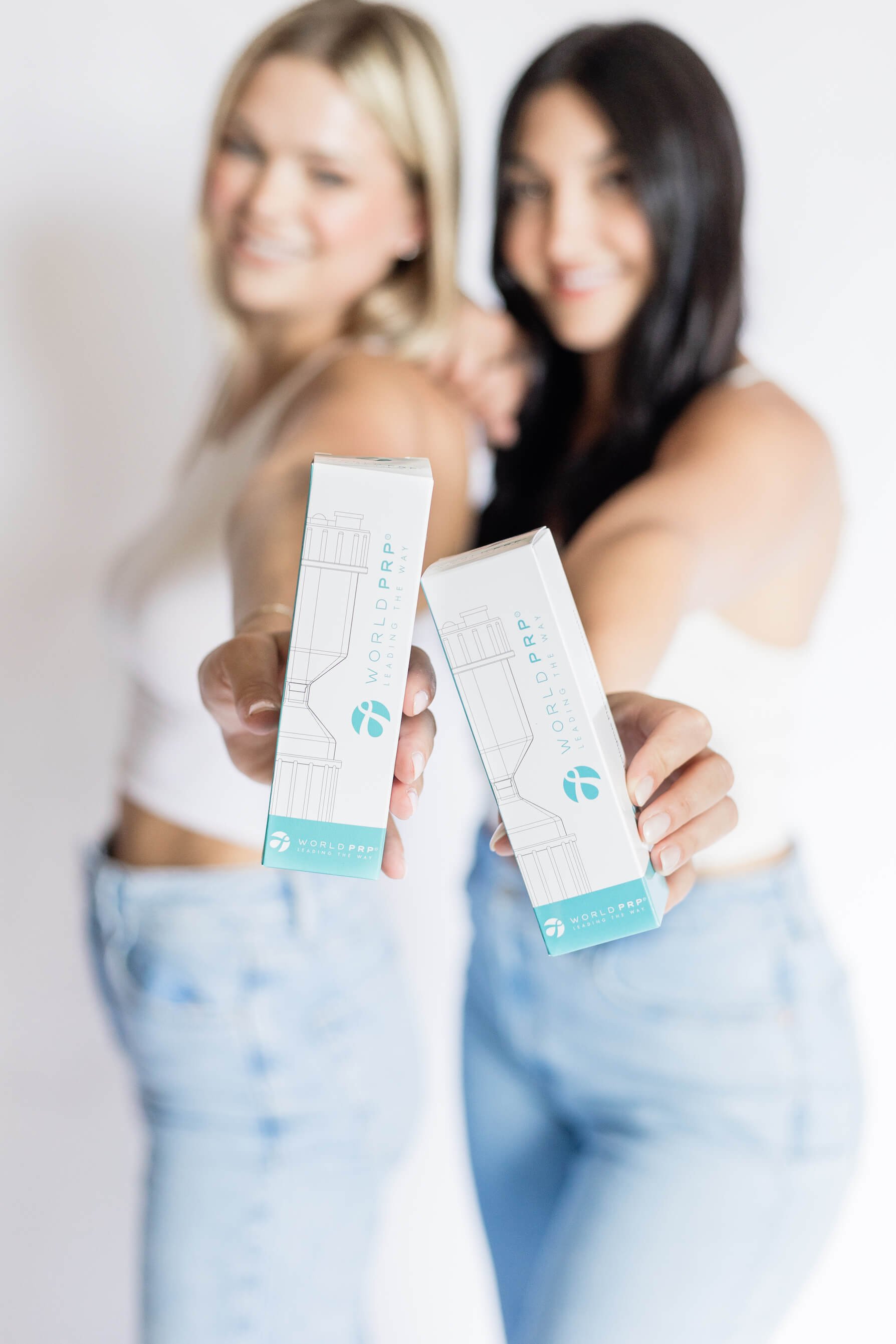 Two women holding PRP product boxes, smiling and wearing light-colored tops and jeans, with a bright background.