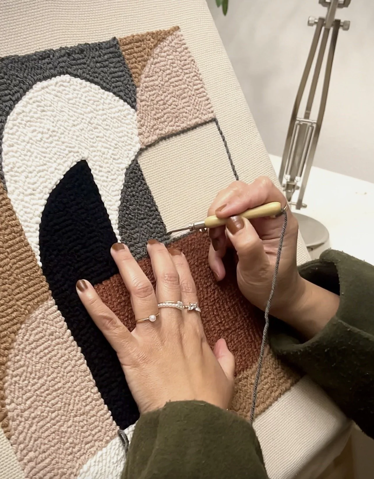 Person creating a geometric textured art piece with black, white, gray, pink, and brown yarns on a canvas, using a felting needle in an indoor setting.