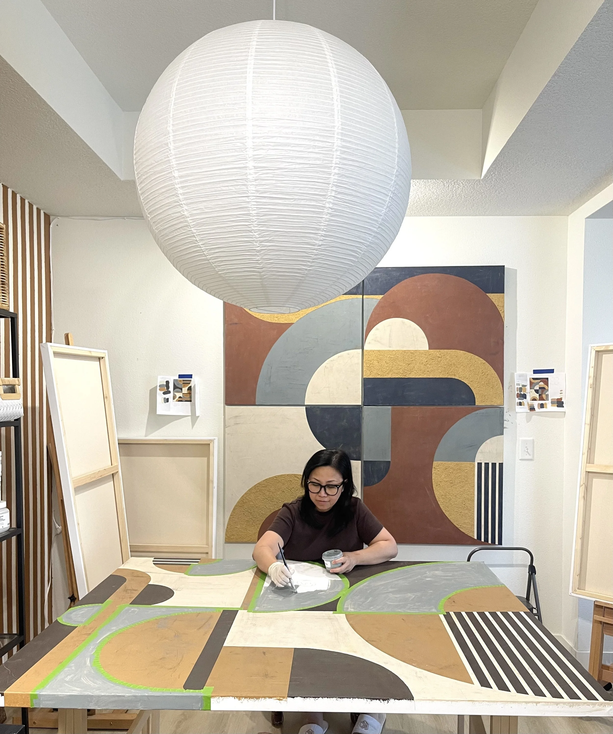 A woman with glasses is painting a colorful geometric mural on a large table in an art studio. The mural features patterns with circles, lines, and blocks of various colors including gray, green, black, and brown. Behind her are abstract paintings and art supplies.