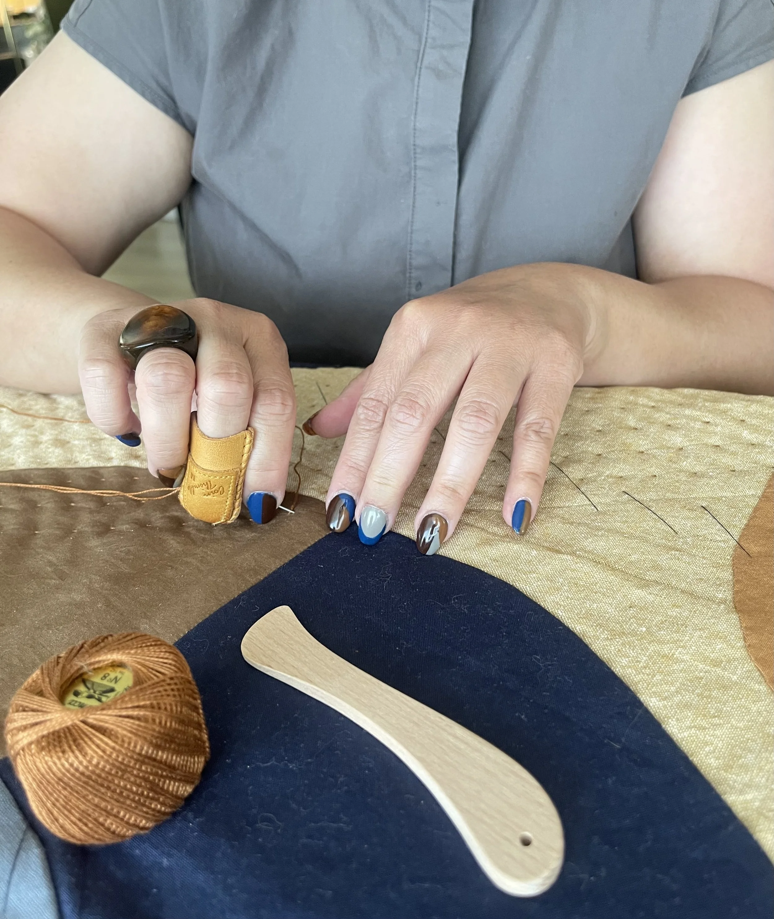 Close-up of a person's hands working on a sewing project, with a ball of bronze-colored thread, a wooden spoon, and a wooden-handled tool on a fabric surface. The person is wearing a grey shirt and has painted nails.