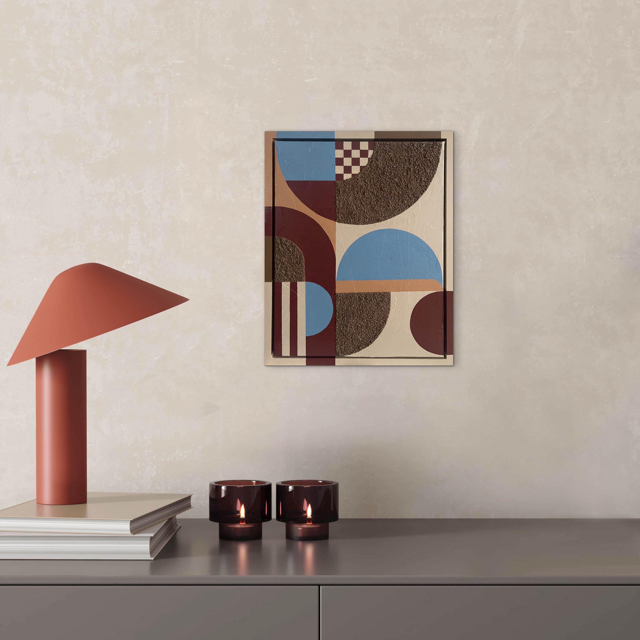 A minimalist interior scene with a white wall, a light-colored rectangular table, a pink desk lamp on the left, a stack of large books on the table, two lit black candles with flames, a framed abstract artwork on the wall, and a textured art piece with geometric patterns and curved shapes in earthy tones and blues.