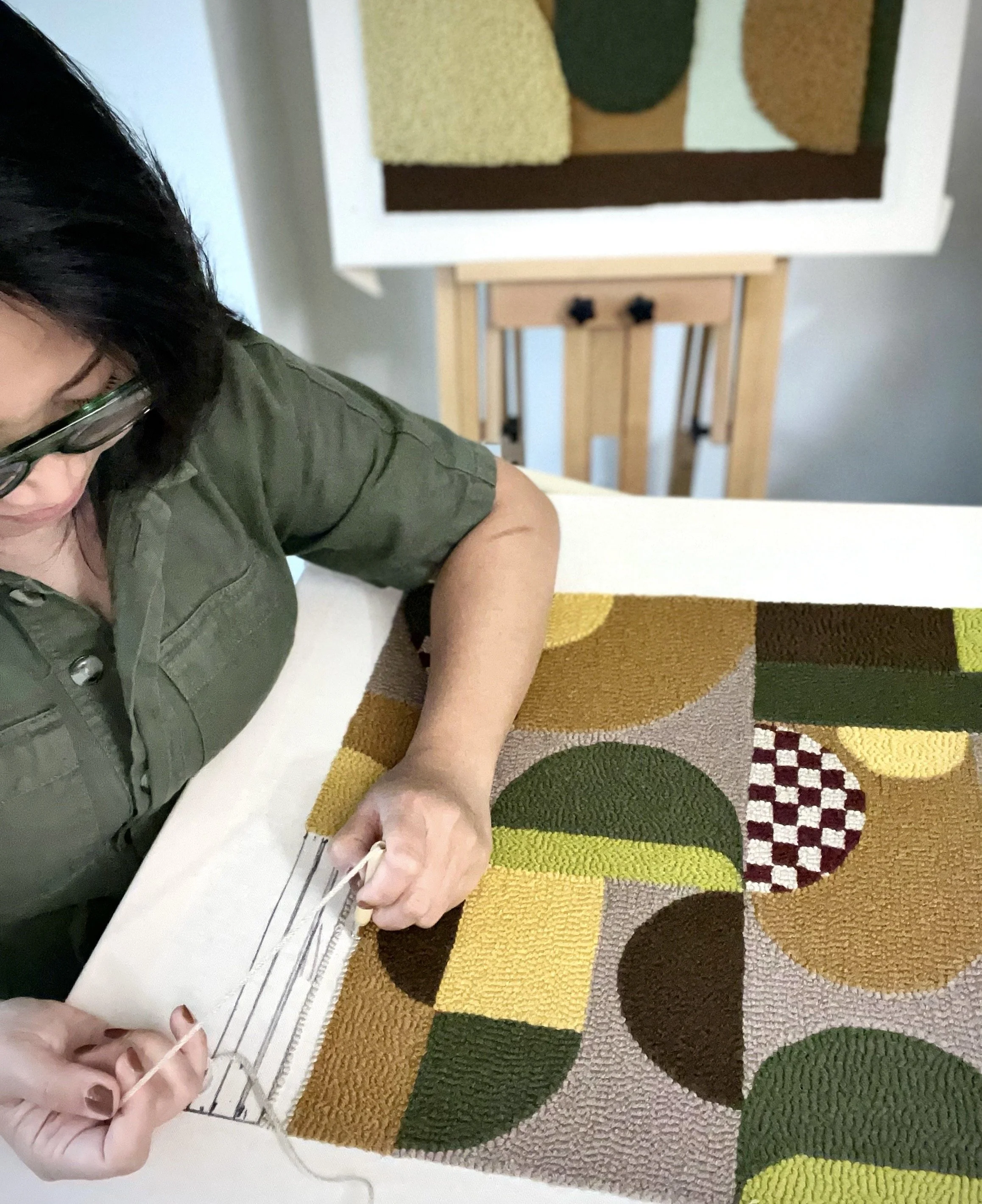 Close-up of a person with black hair, wearing glasses and a green shirt, working on a colorful, patterned woven piece, likely a rug, with geometric shapes in shades of yellow, orange, green, brown, and black.