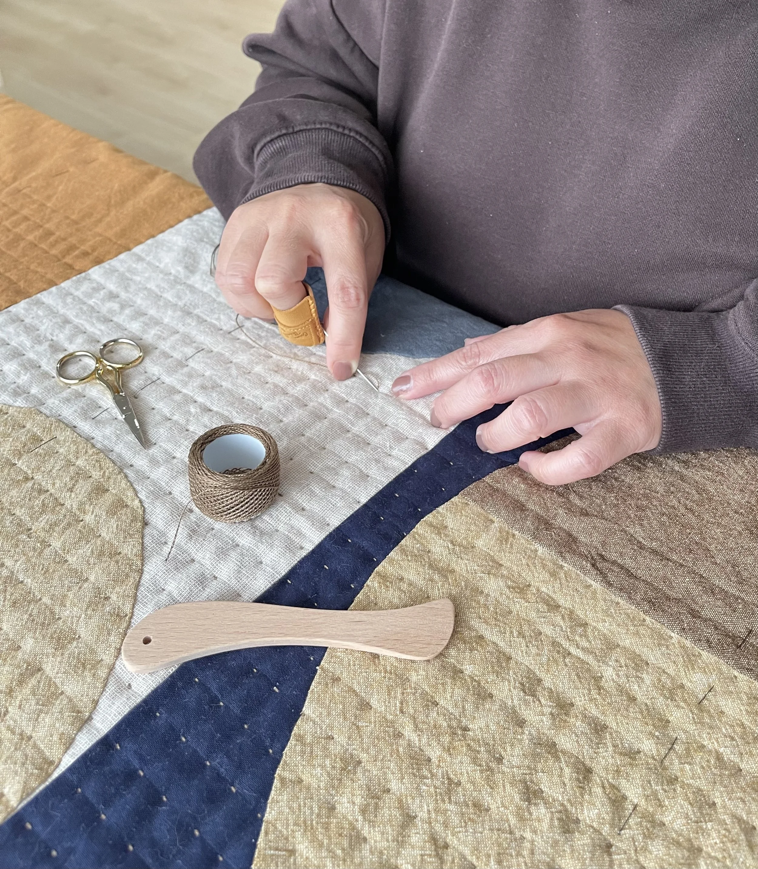 A person sewing on fabric with a needle and thread, with sewing scissors, a spool of thread, and a wooden tool on a table.