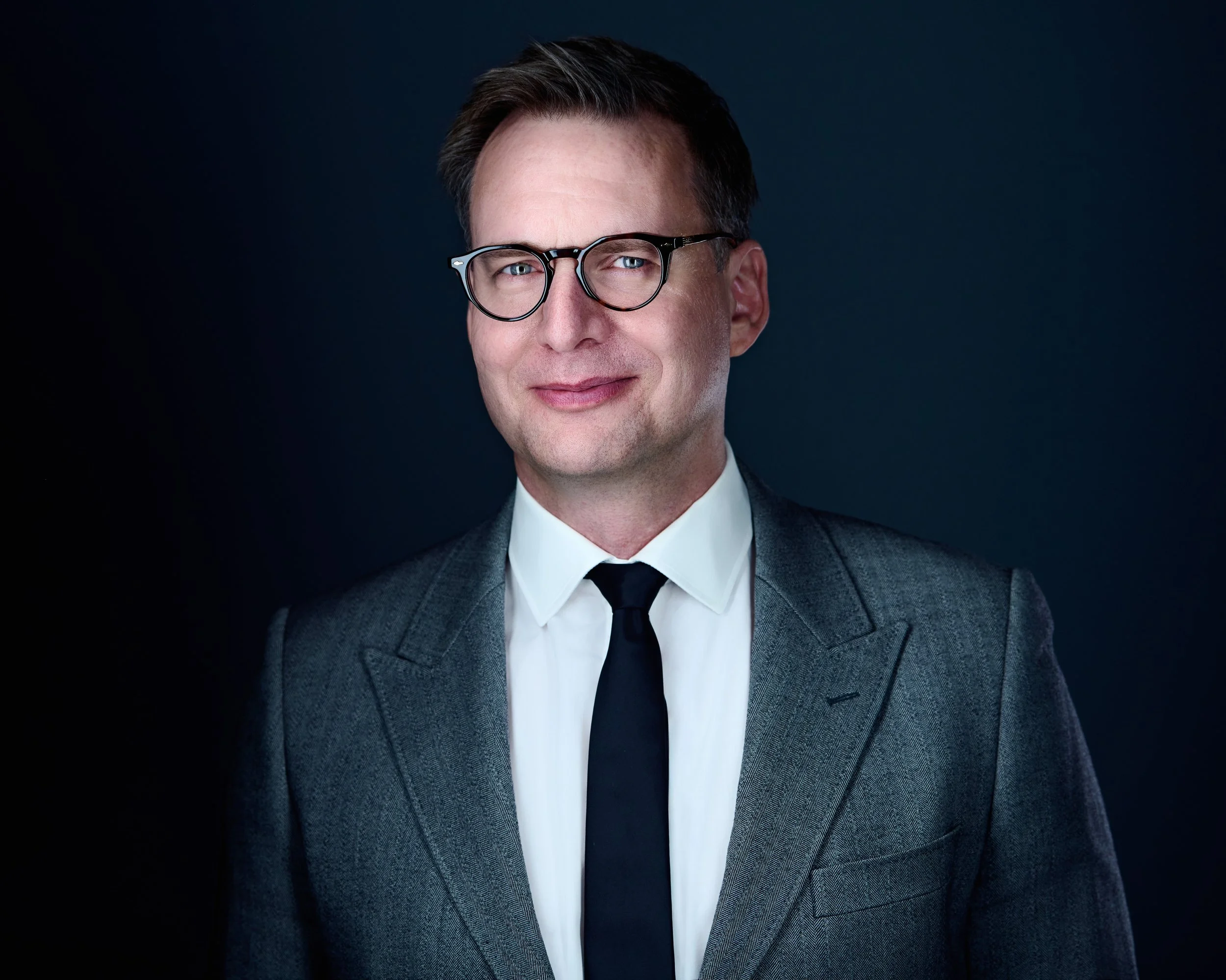 Portrait of a man wearing glasses, a gray suit, white shirt, and black tie, against a dark background.