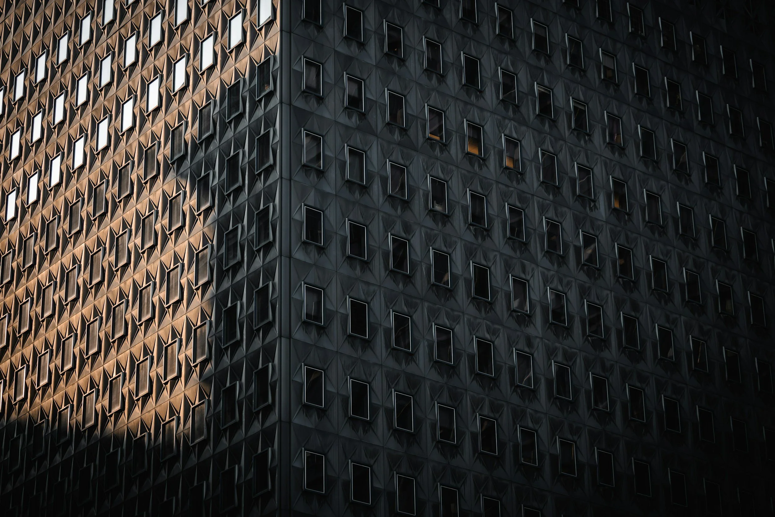 Close-up of a modern black building with numerous small, rectangular windows, some reflecting light and some dark, creating a geometric pattern.