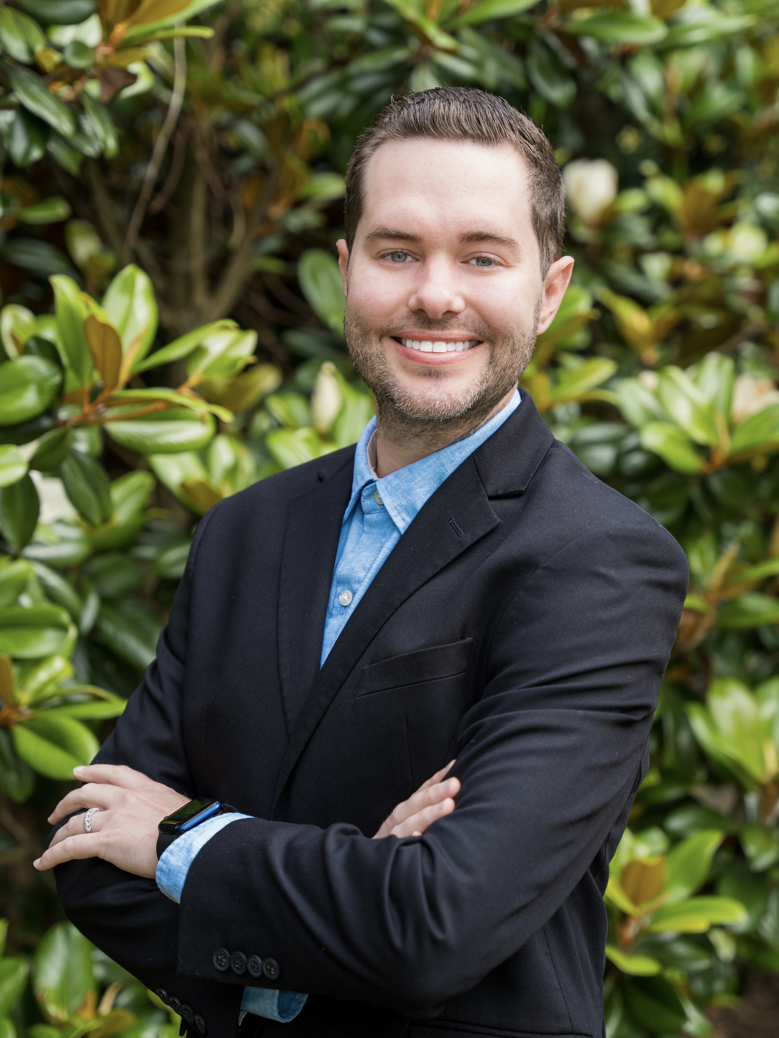 A smiling man with brown hair and a beard, wearing a black blazer and light blue button-up shirt, standing with arms crossed in front of green leafy bushes.