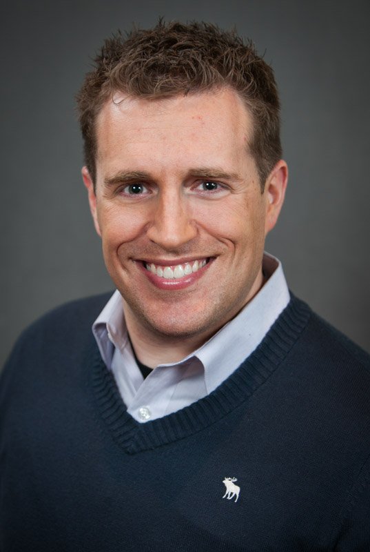 Headshot of a smiling man with short, curly brown hair, wearing a white collared shirt under a dark blue sweater with a moose logo, against a gray background.