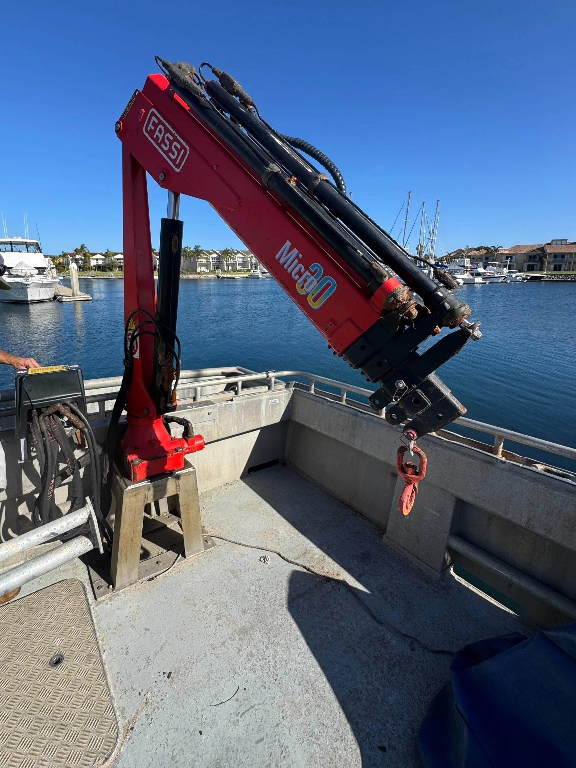 A red Fassi crane mounted on a boat with a hook hanging down, positioned near the water with boats and houses in the background.
