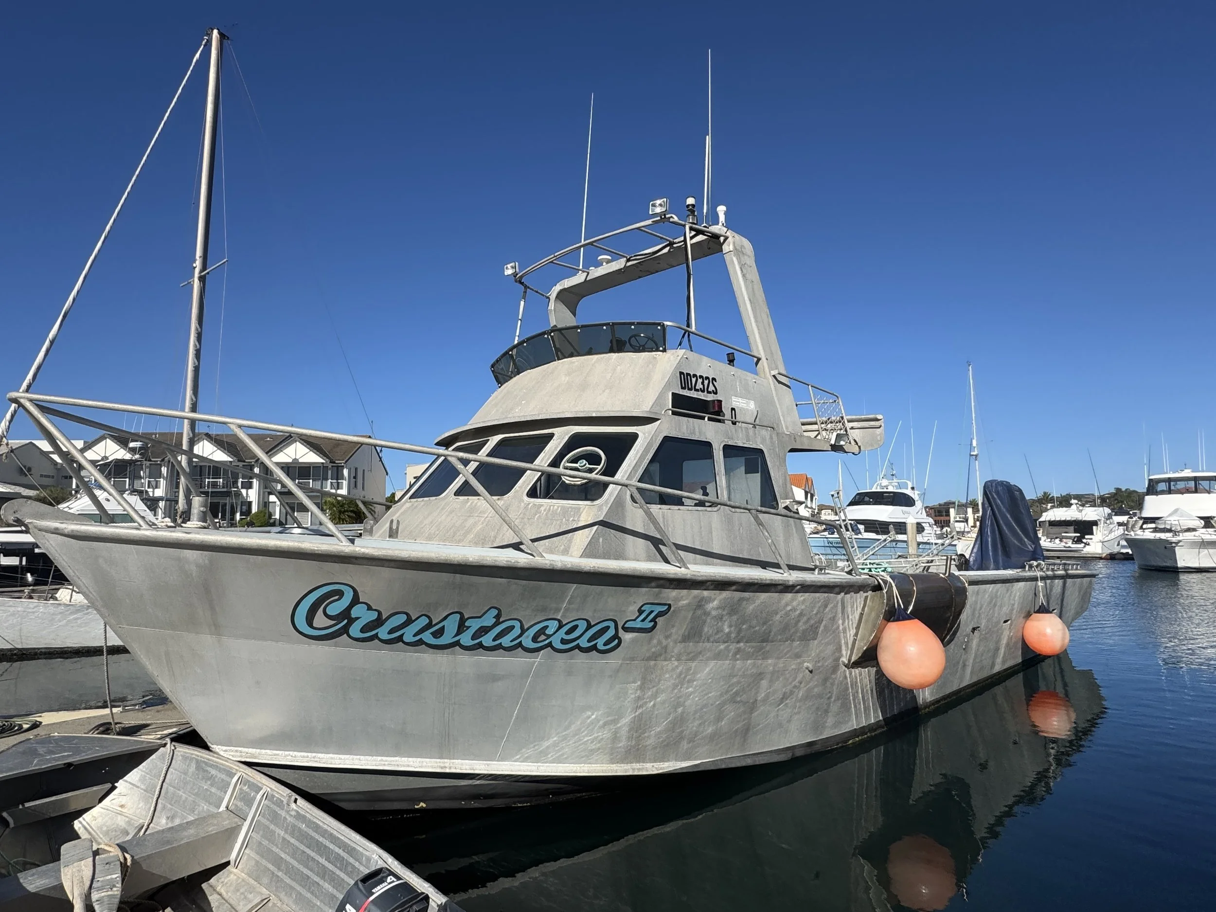 A large fishing or recreational boat named Crustacean II docked at a marina under a clear blue sky, with other boats and marina buildings in the background.