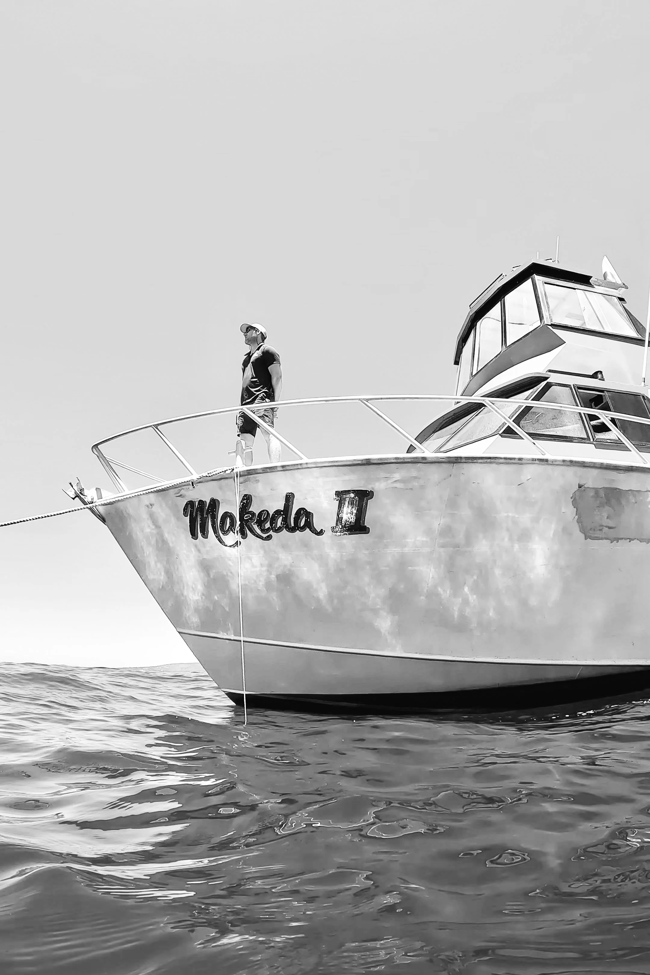 A man standing on the bow of a boat named 'Makeda II' floating on water, with a clear sky in the background.