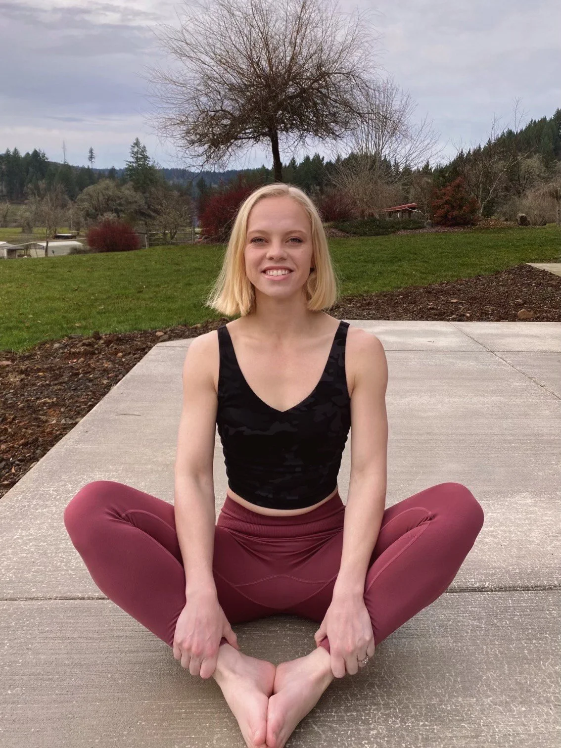 A young woman with blonde hair sitting cross-legged on a concrete patio outdoors, with a grassy area, trees, and mountains in the background.