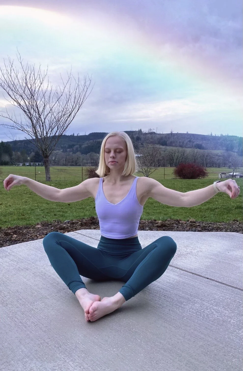 A woman practicing yoga or meditation outdoors on a mat, sitting cross-legged with arms extended and eyes closed, with a scenic rural landscape and cloudy sky in the background.