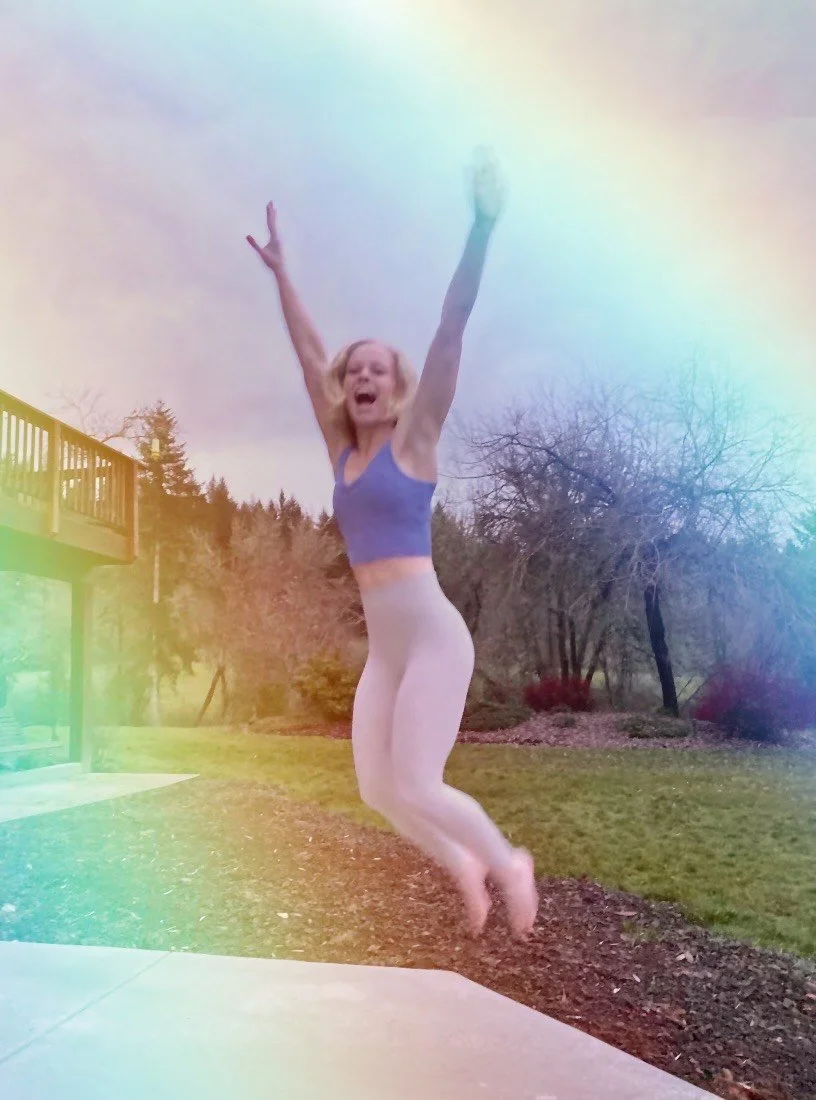A woman in a blue sports bra and white leggings jumping into the air with arms raised, outdoors near a backyard with trees and a deck.
