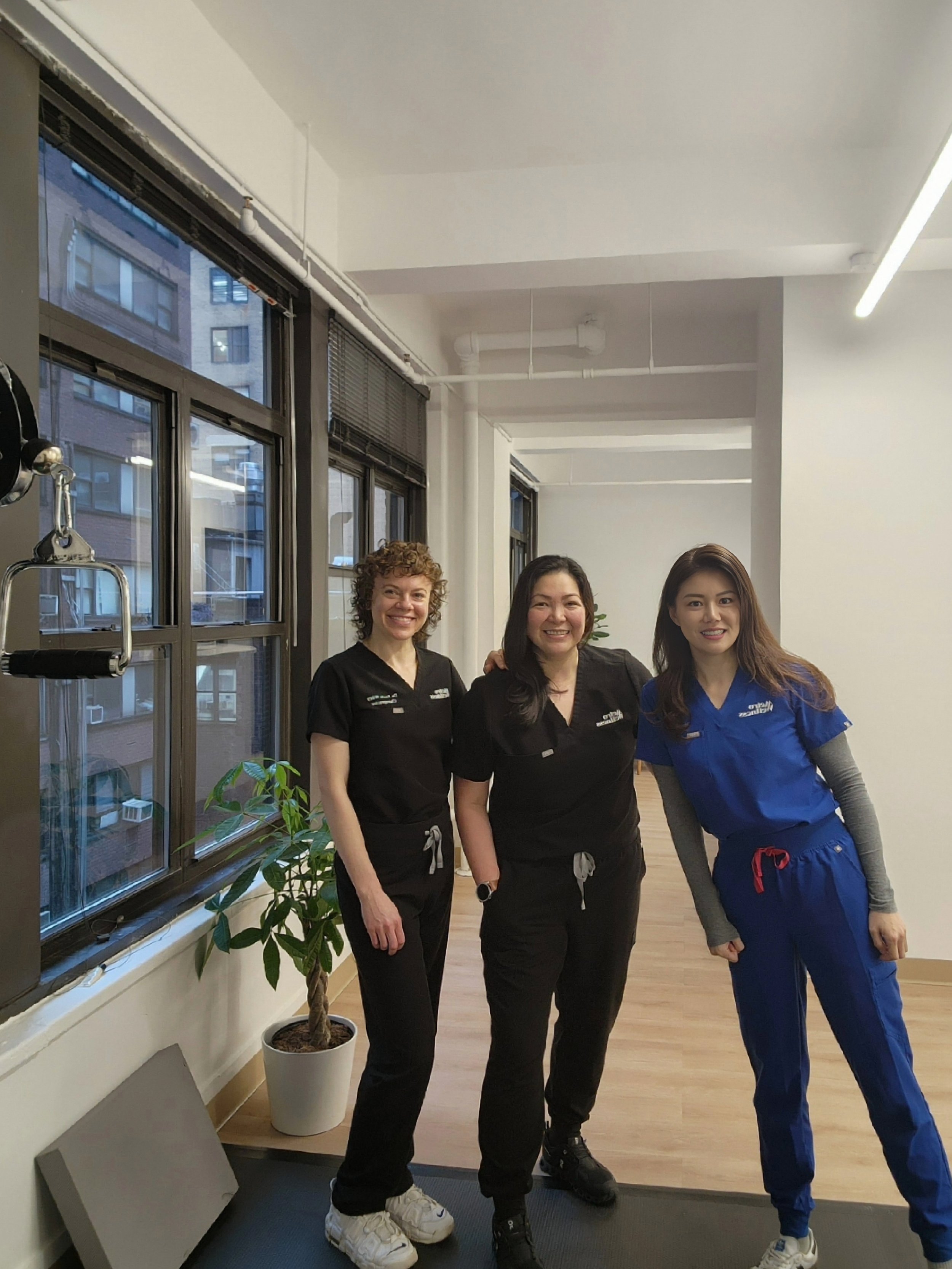 Three women in medical scrubs standing inside a room near large windows.
