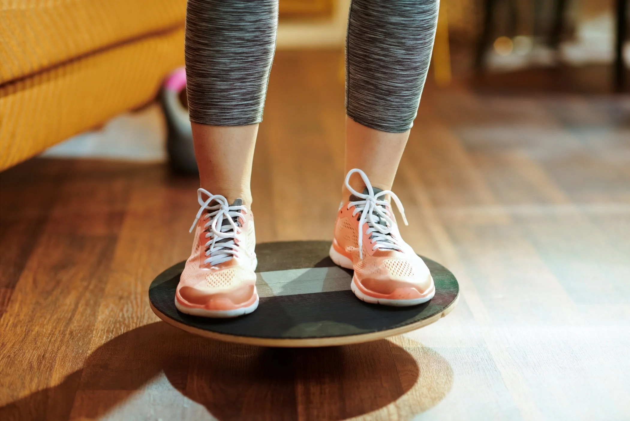 Person standing on a balance board indoors, wearing gray leggings and peach running shoes.