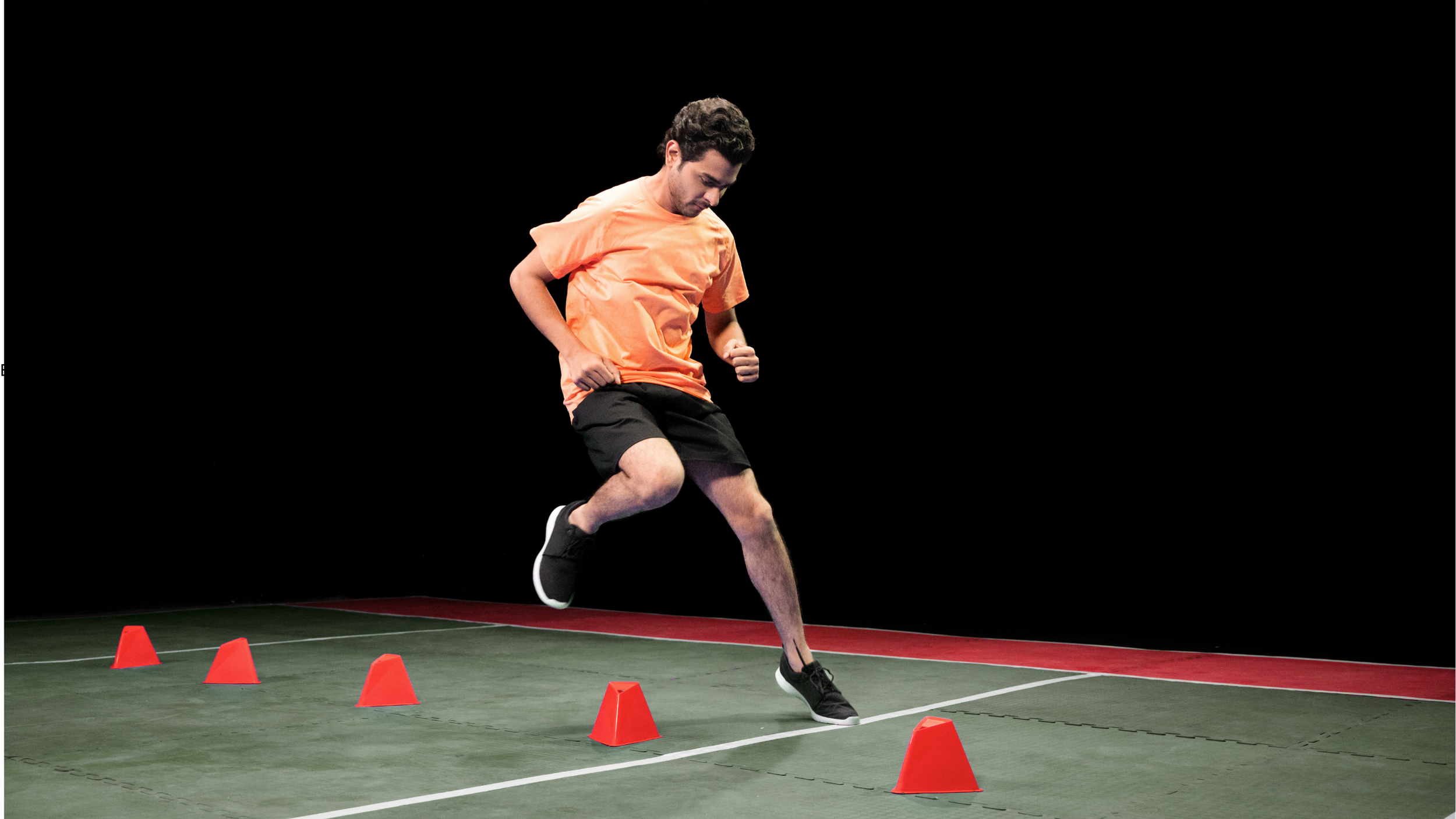 Man in orange shirt and black shorts doing agility drill with orange cones on indoor court.
