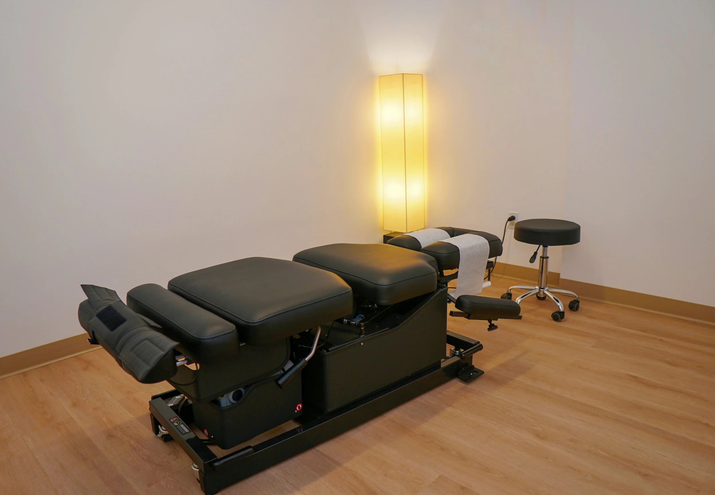 Massage table with black cushions in a room with wooden floors, a tall yellow floor lamp, and a small black stool.