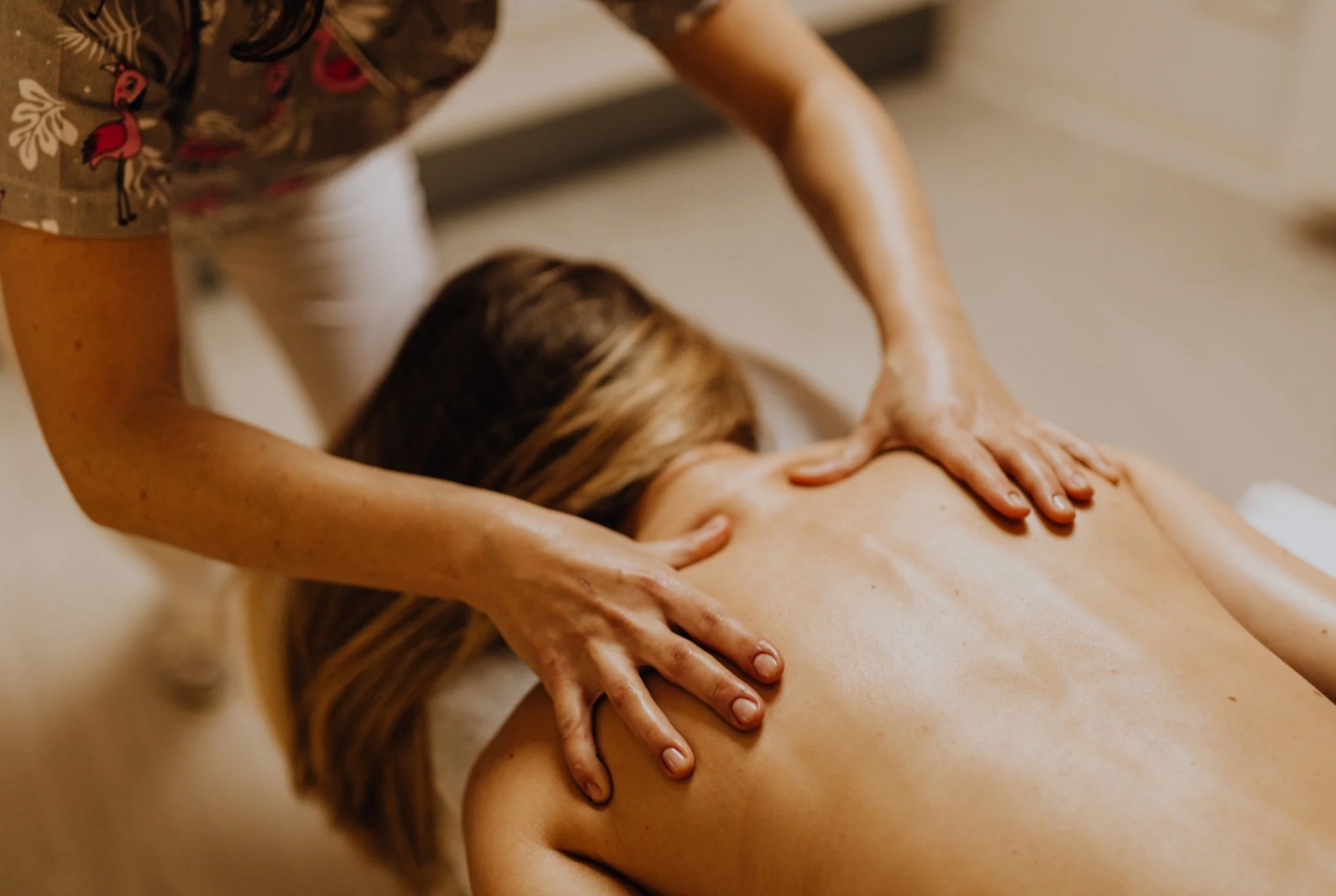 A person receiving a back massage, lying face down on a massage table. The massage therapist is using both hands to apply pressure to the person's back.