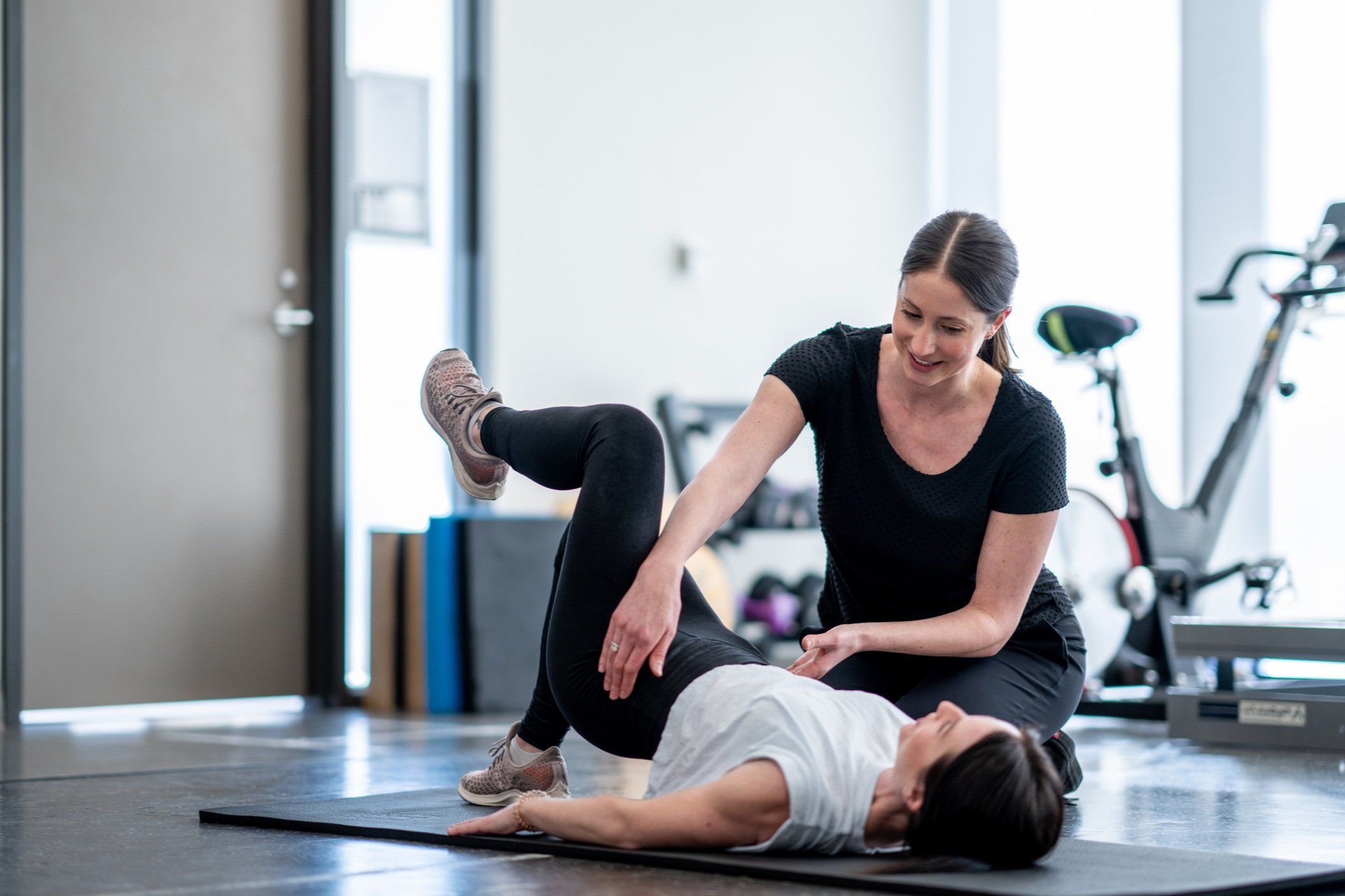 A personal trainer assisting a woman exercising on a yoga mat in a gym with exercise bikes in the background.