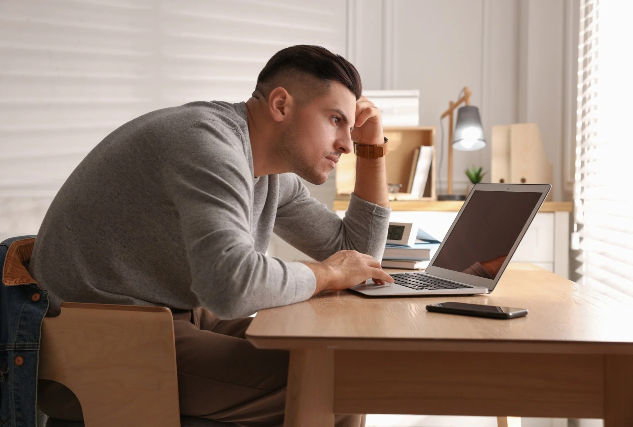 A man sitting at a desk looking at a laptop with a pensive expression, resting his head on his hand.