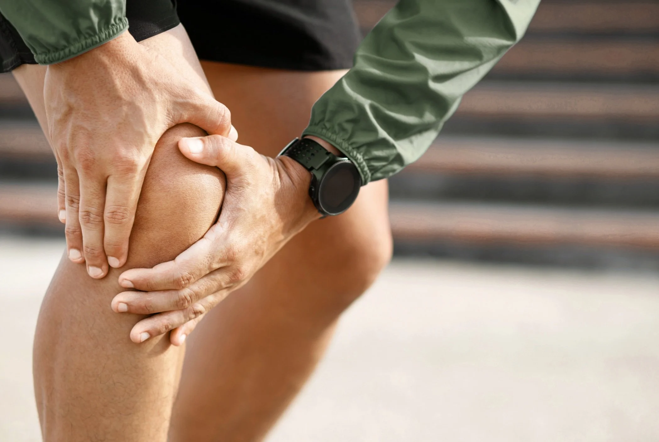A person holding their knee in pain, outdoors, wearing athletic clothing and a smartwatch.